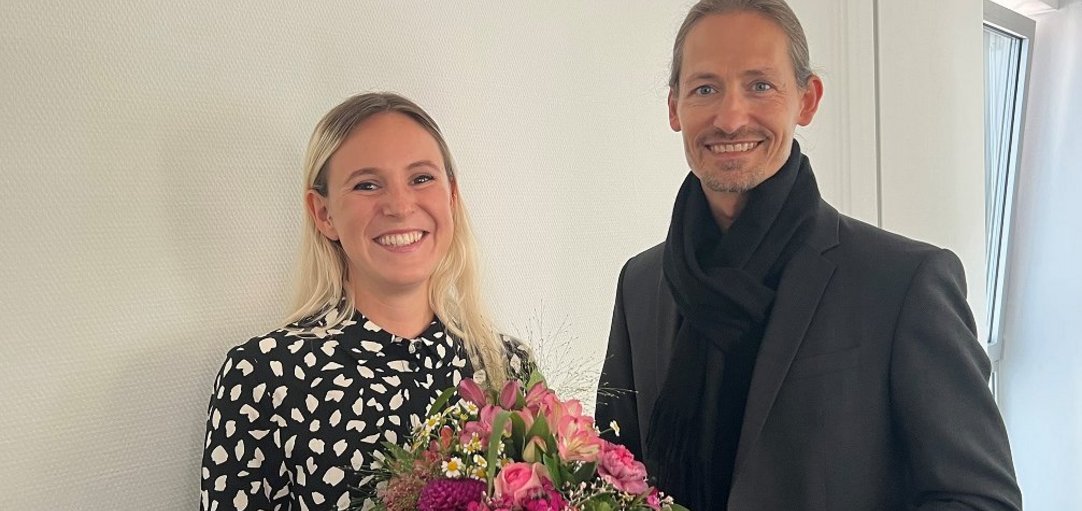 Auf dem Bild stehen ein Mann und eine Frau nebeneinander. Die Frau hält einen Blumenstrauß in der Hand. 