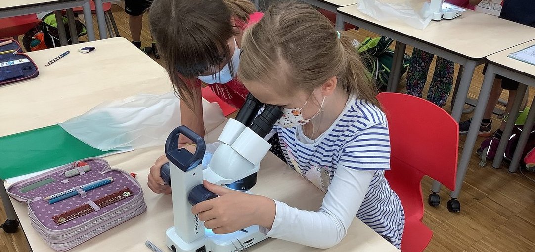 Zwei Grundschülerinnen sitzten im Klassenraum und betrachten etwas durch ein Mikroskop. 