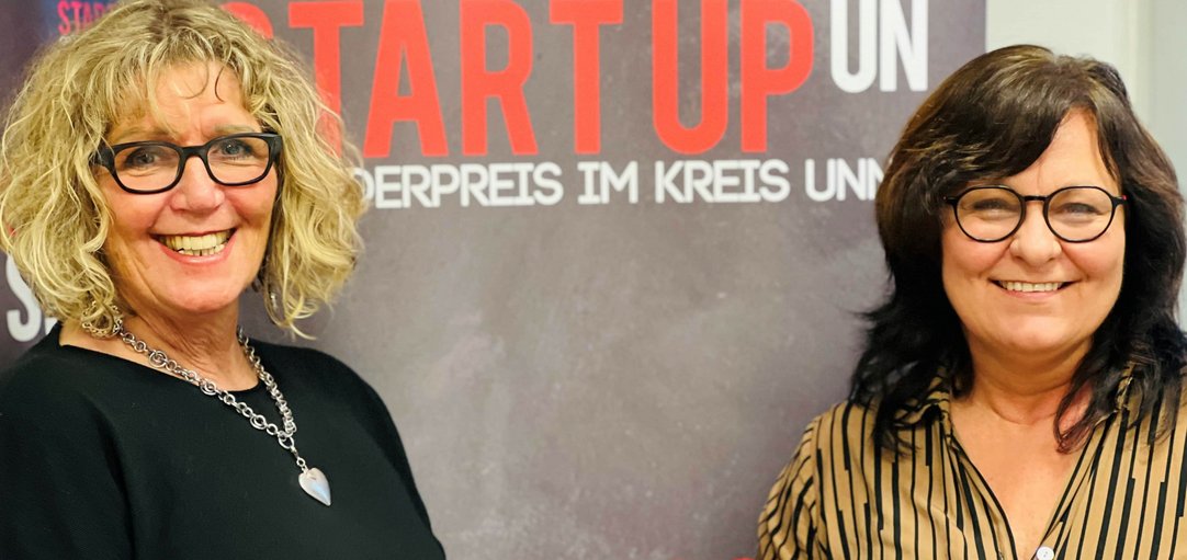 Die Gründerin und die Gründungsberaterin stehen lächelnd vor einem Roll-Up mit Titel "StartupUN".