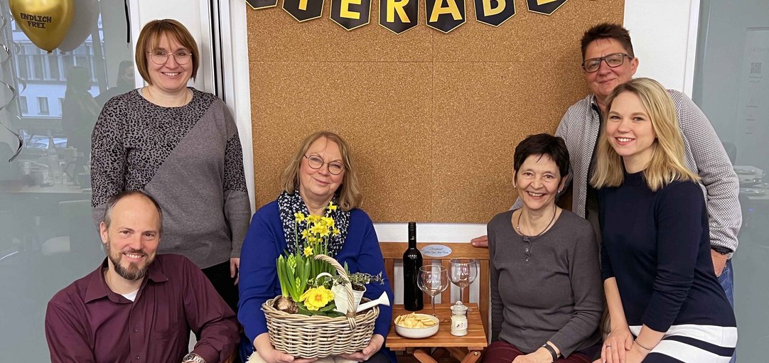 Teamfoto, sechs Personen lachend mit Blumen, Luftballons und dem Schild Feierabend.